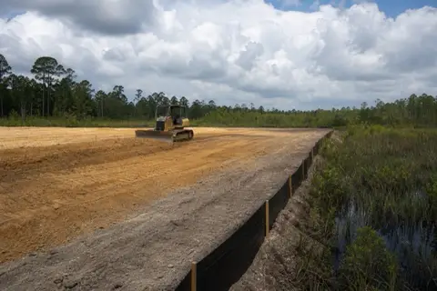 Small dozer leveling a homesite pad with grade stakes and silt fence along the low wet edge